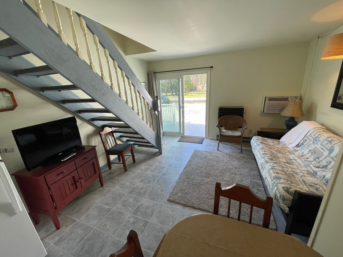 A living area is depicted, featuring a sleeper sofa with a patterned cover and a wooden coffee table. A flat-screen television is positioned on an entertainment unit. A sliding glass door leads to an outdoor space, while stairs ascend to the upper level.