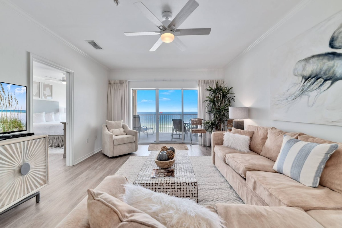 A spacious living area features a light-colored sofa and armchair, complemented by a textured coffee table. Large sliding glass doors provide access to a balcony with views of the Gulf. Natural light floods the room, highlighting coastal decor and neutral tones.