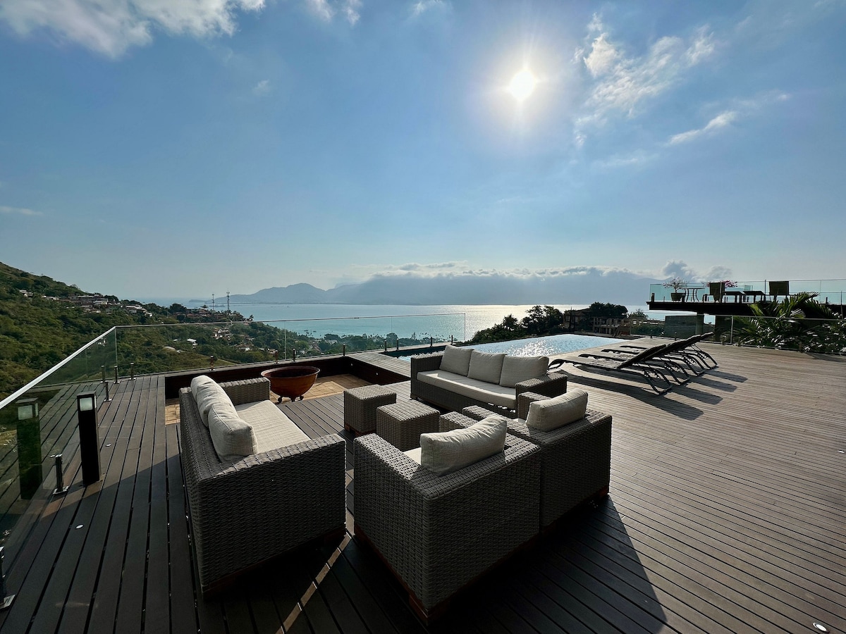 An outdoor lounge area features comfortable seating arranged around a central table, overlooking a stunning view of the sea. A heated infinity pool extends in the background, with sun loungers positioned for relaxation under the bright sun.