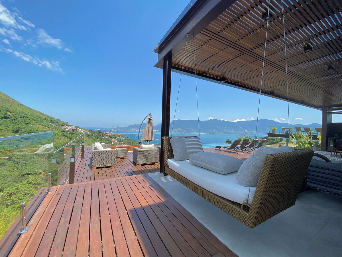 An outdoor lounge area features comfortable seating arrangements, with a swinging sofa overlooking a panoramic view of the ocean and mountains. The deck is constructed of wooden planks, complemented by glass railings that enhance the scenic environment.
