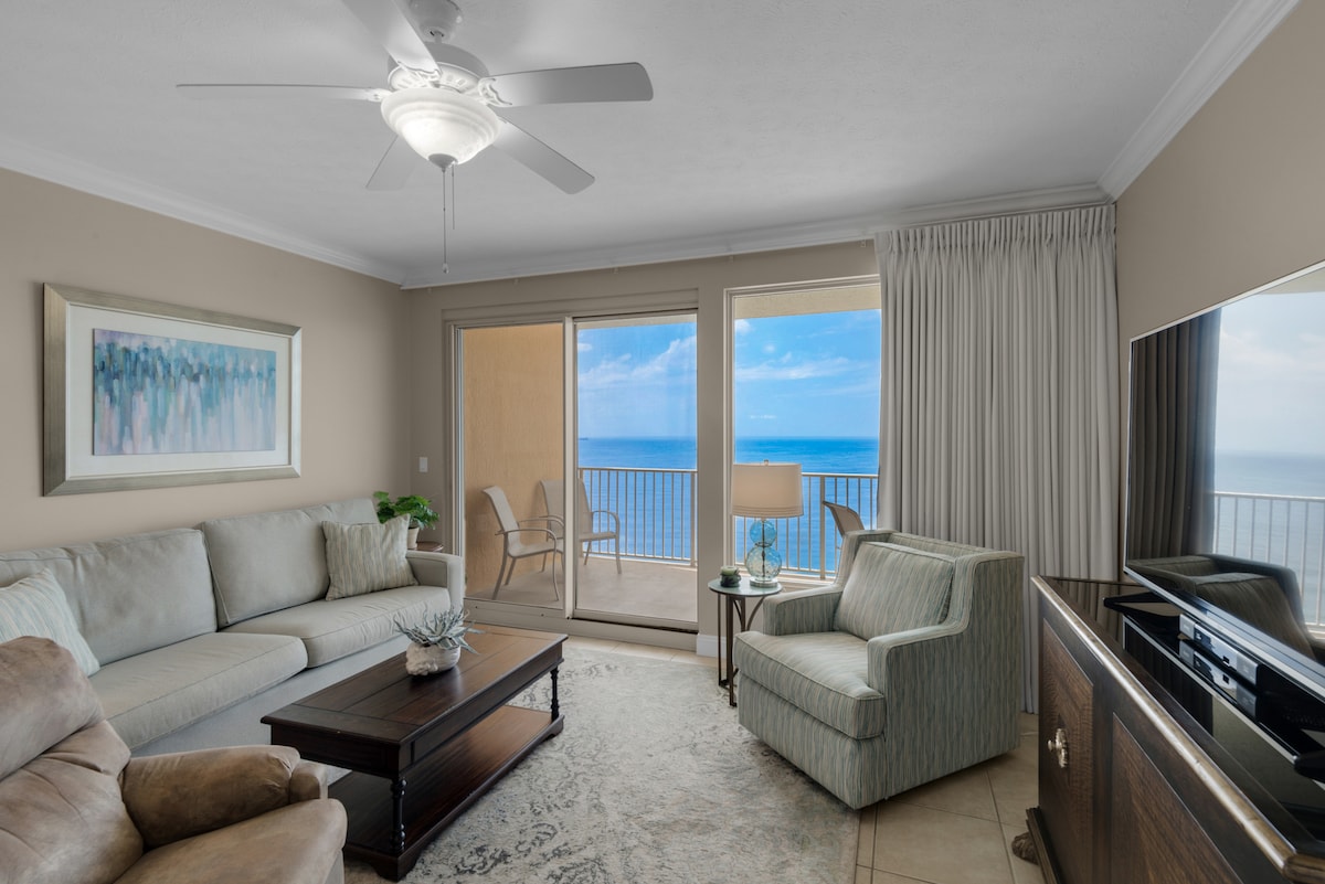 The living area features a light-colored sofa and a plush armchair, facing a dark wooden coffee table. A flat-screen TV is positioned across from the seating. Large sliding glass doors open to a private balcony, revealing expansive views of the ocean.