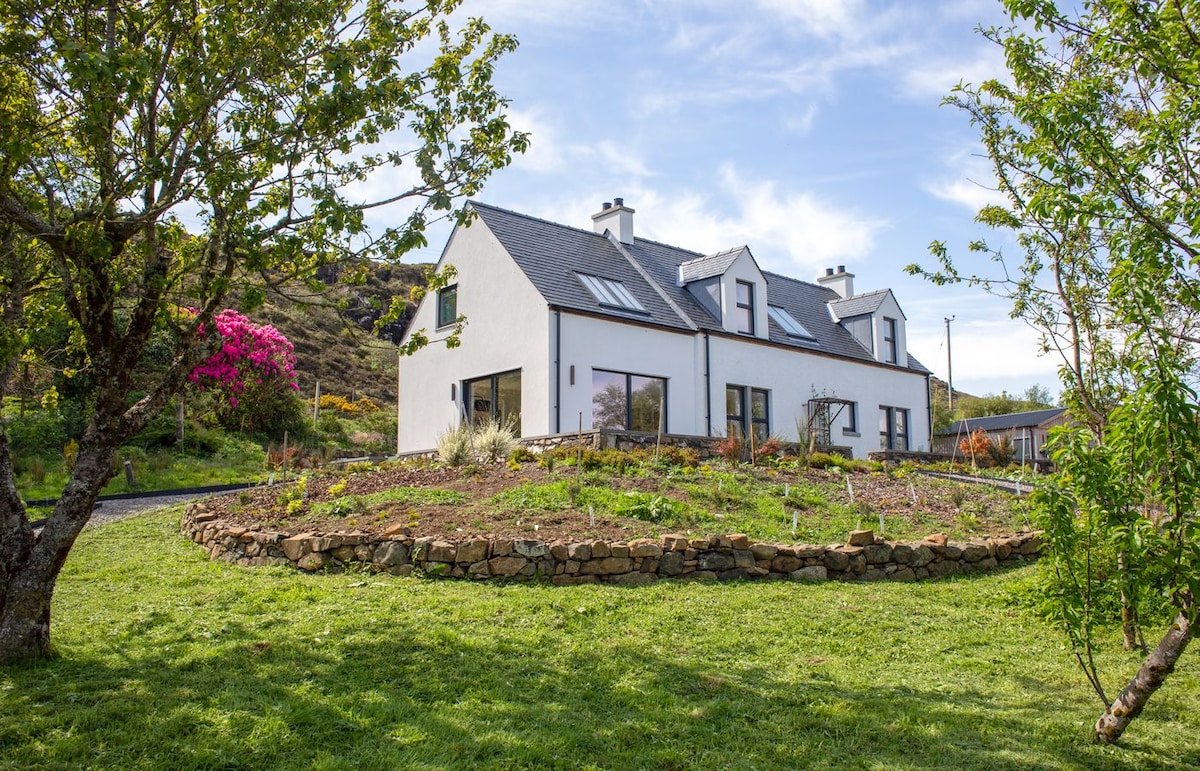 A modern white cottage is framed by a landscaped garden, featuring a circular arrangement of stone borders. Surrounding trees and blooming flowers add color, while the blue sky above contributes to the serene atmosphere of the property.