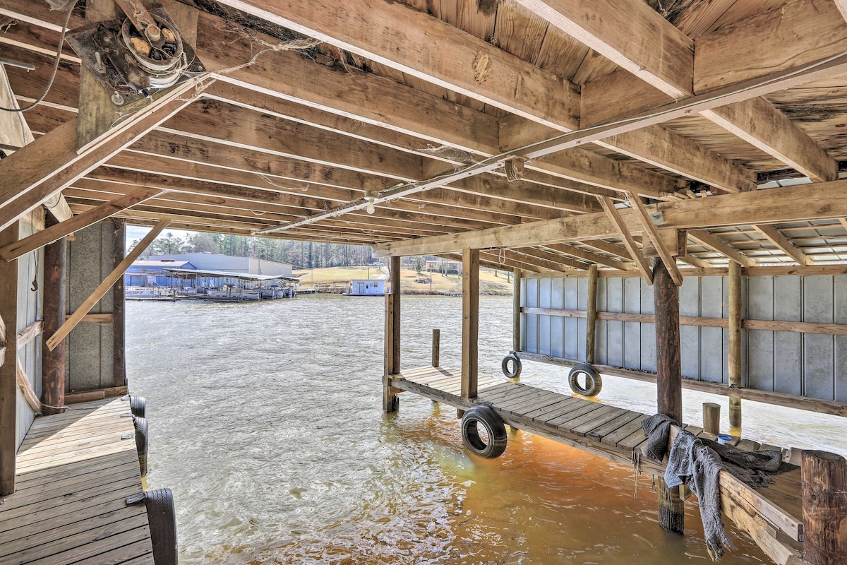 A covered boat dock features wooden beams overhead and wooden planks underfoot. Water ripples gently below, with tires hanging from the structure. Visible on the horizon are signs of neighboring docks and shoreline, surrounded by trees. Natural light filters through the open areas.