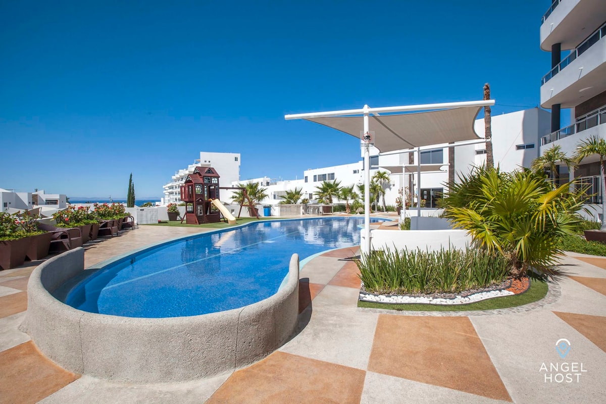 The image captures a spacious pool area surrounded by well-manicured landscaping. A winding pool features clear blue water and is complemented by lounge chairs and palm trees. The sun is shining under a clear blue sky, creating a bright and inviting atmosphere.