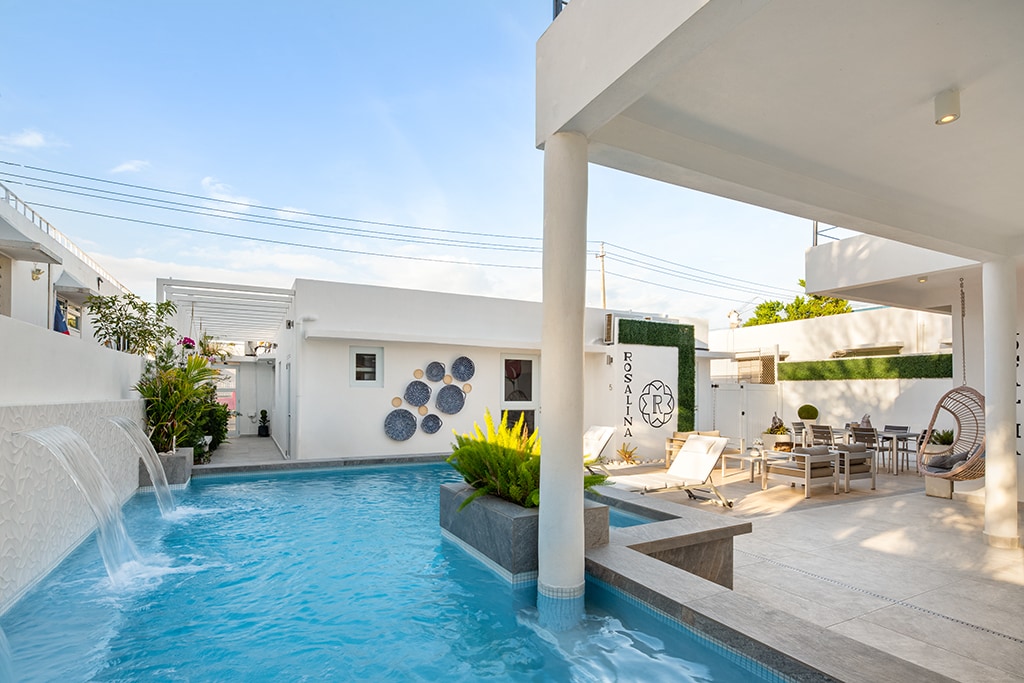 A shared pool area is presented, featuring clear blue water with gentle fountains. Surrounding lounge chairs are arranged neatly, accompanied by a shaded seating area. The space is framed by modern architectural elements and green landscaping, creating a tranquil environment for relaxation.