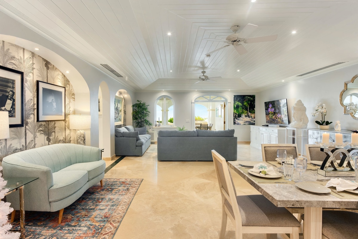 An open-concept living and dining area features a light color palette with stylish furniture, including a teal sofa and a wooden dining table. Arched windows offer views of the Caribbean Sea, and soft lighting enhances the tranquil atmosphere.