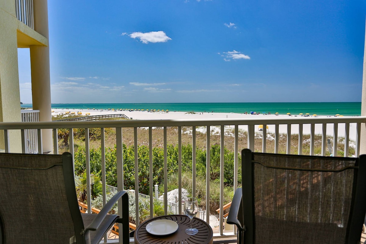 A direct beachfront balcony offers a spacious seating area with two chairs and a small table. Views of the Gulf of Mexico and the sandy beach are visible, with a clear blue sky enhancing the serene environment.