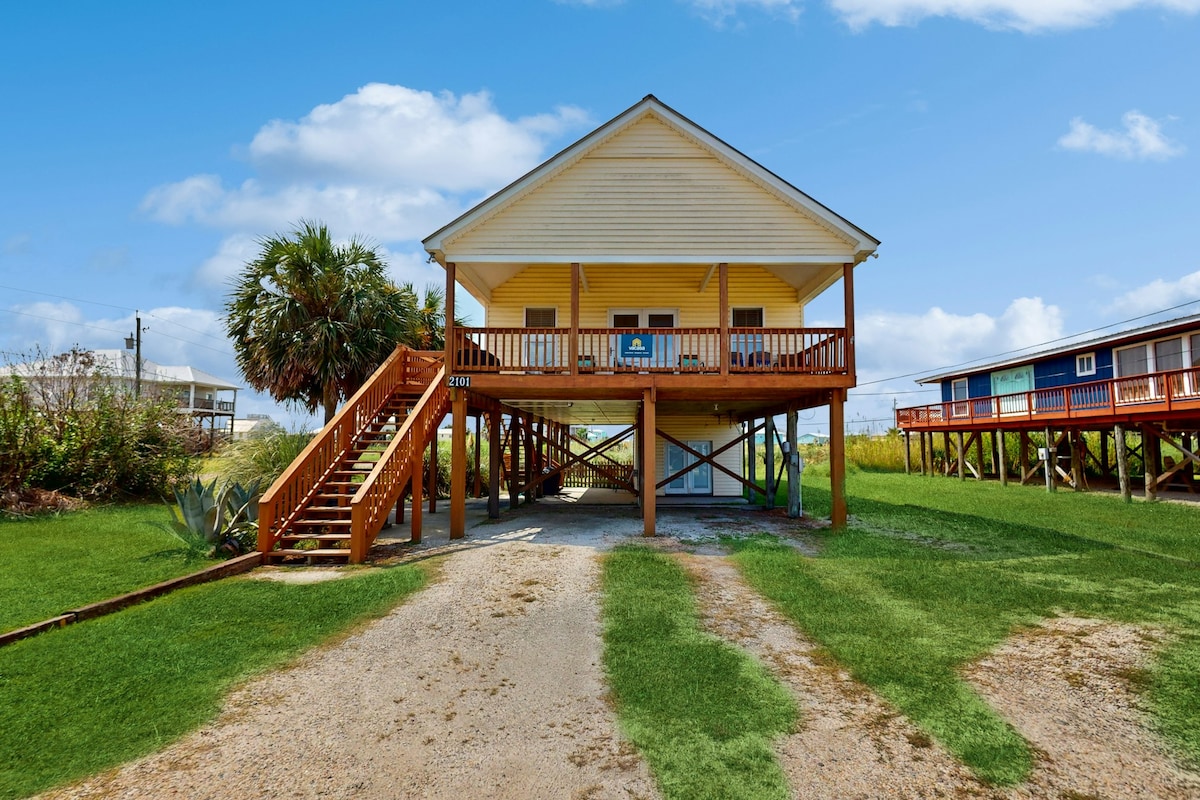 A two-story house with a yellow exterior and a welcoming front porch is shown. Sturdy wooden steps lead up to the porch, surrounded by lush green grass and palm trees. The blue sky serves as a backdrop, creating an inviting atmosphere.