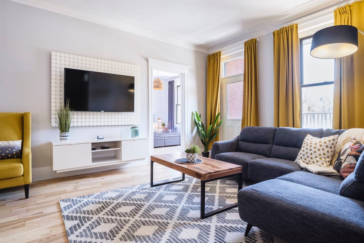 The spacious living area features a large corner sofa, complemented by a wooden coffee table. A flat-screen TV is mounted on a light-colored wall. Bright curtains frame the windows, allowing natural light to enhance the comfortable ambiance. A decorative plant adds a touch of greenery.