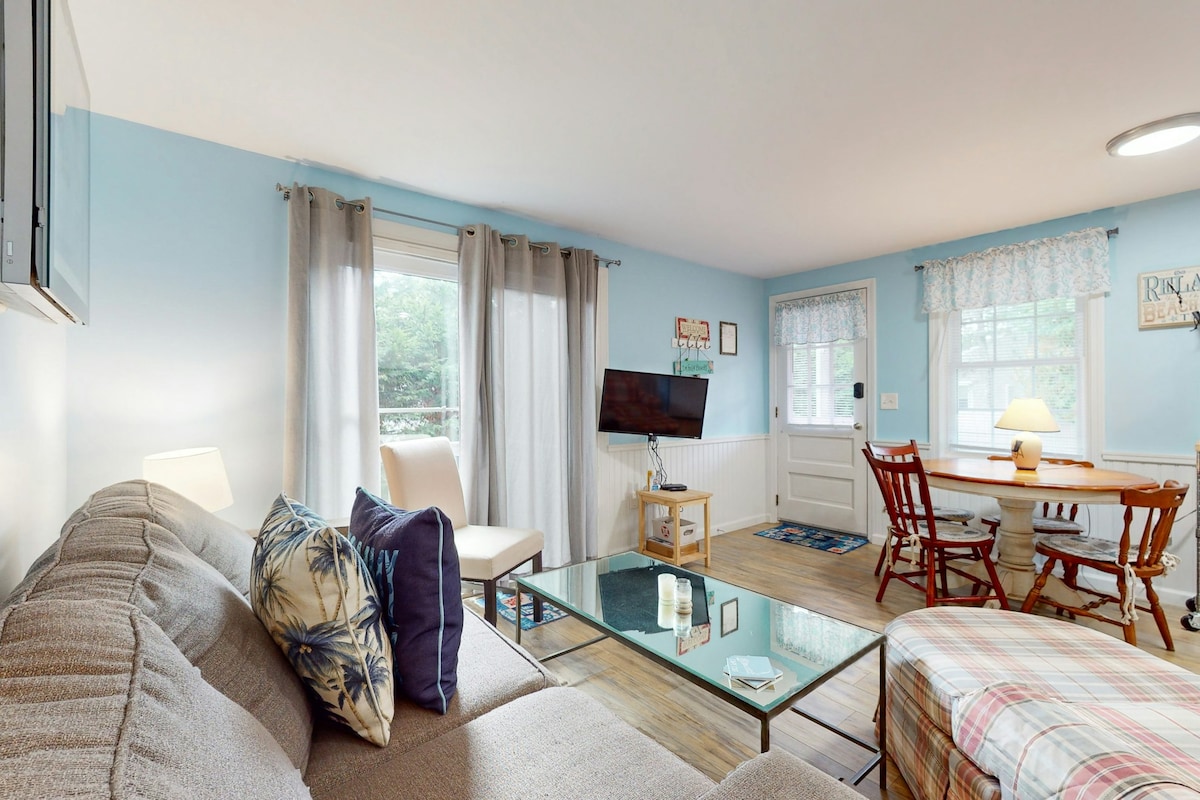 A well-lit living area features a comfortable couch adorned with decorative pillows and a glass coffee table. A TV is mounted on the wall across from the seating. A dining area with a wooden table and chairs is situated near a window, with light filtering through sheer curtains.
