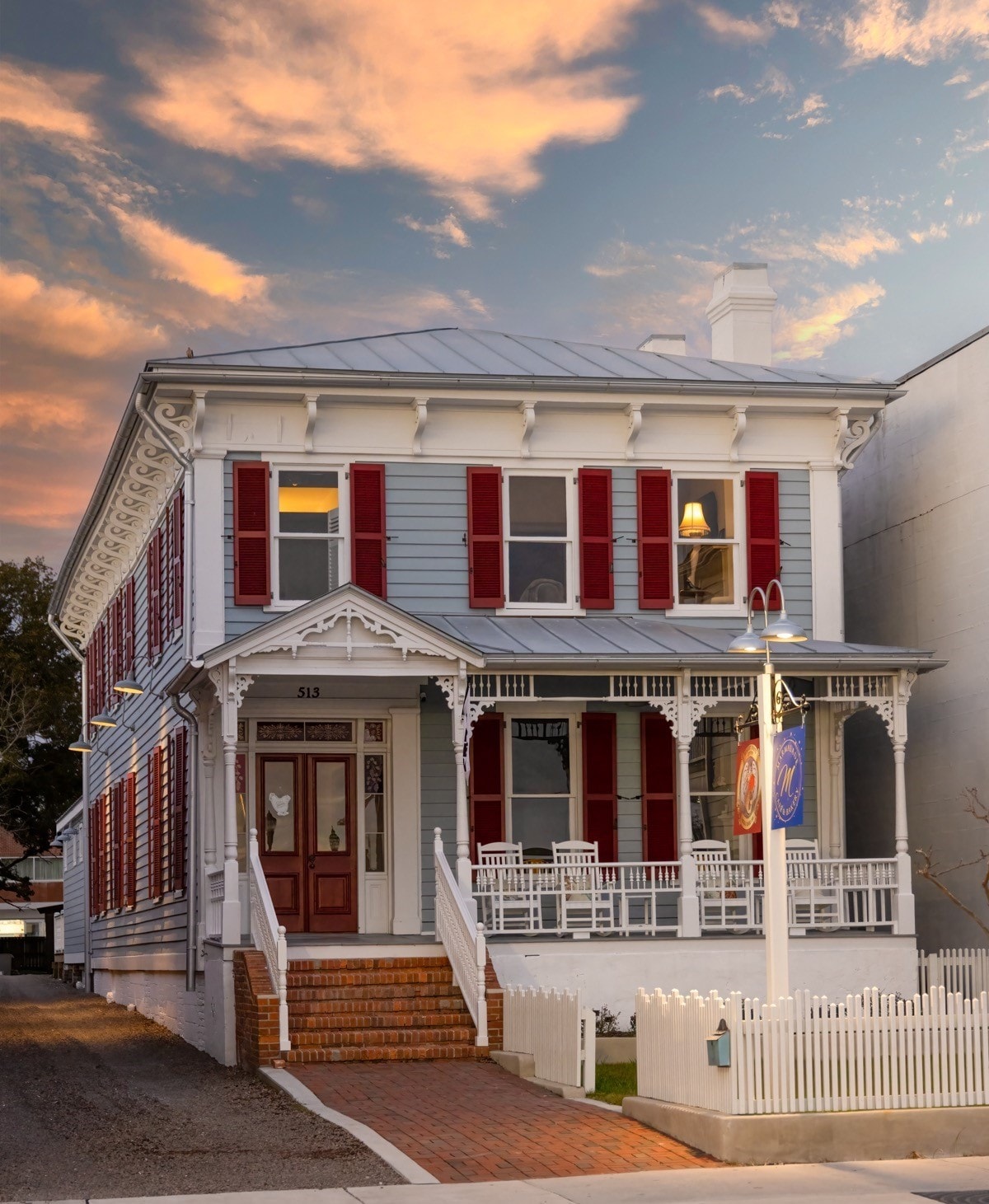 The exterior of a historic building showcases classic architecture with white siding and red shutters. A welcoming front porch features rocking chairs, and steps lead to the entrance. The sky is streaked with soft hues of orange and pink during sunset.
