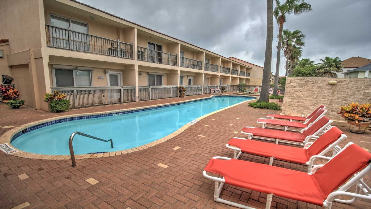 A tranquil pool area is seen, featuring a clear blue swimming pool bordered by lounge chairs. Lush tropical plants surround the space, while a two-story building with balconies frames the pool scene under a cloudy sky.