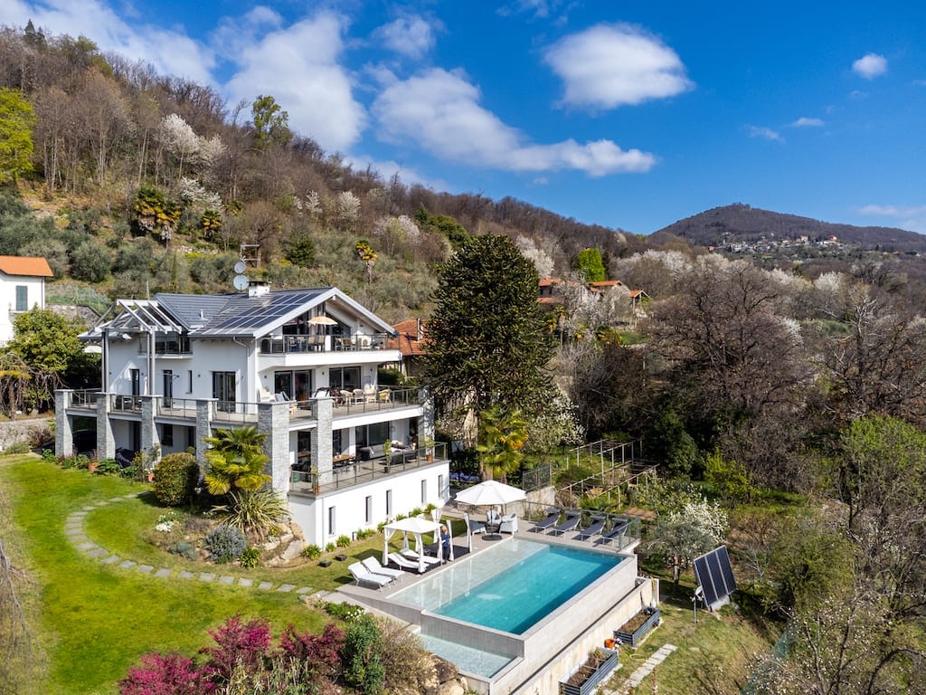 A contemporary villa is nestled against a lush, hilly backdrop. The property features a spacious layout with multiple terraces and an infinity pool surrounded by sun loungers. Verdant gardens with colorful flowering plants enhance the tranquil atmosphere, all under a vibrant blue sky.