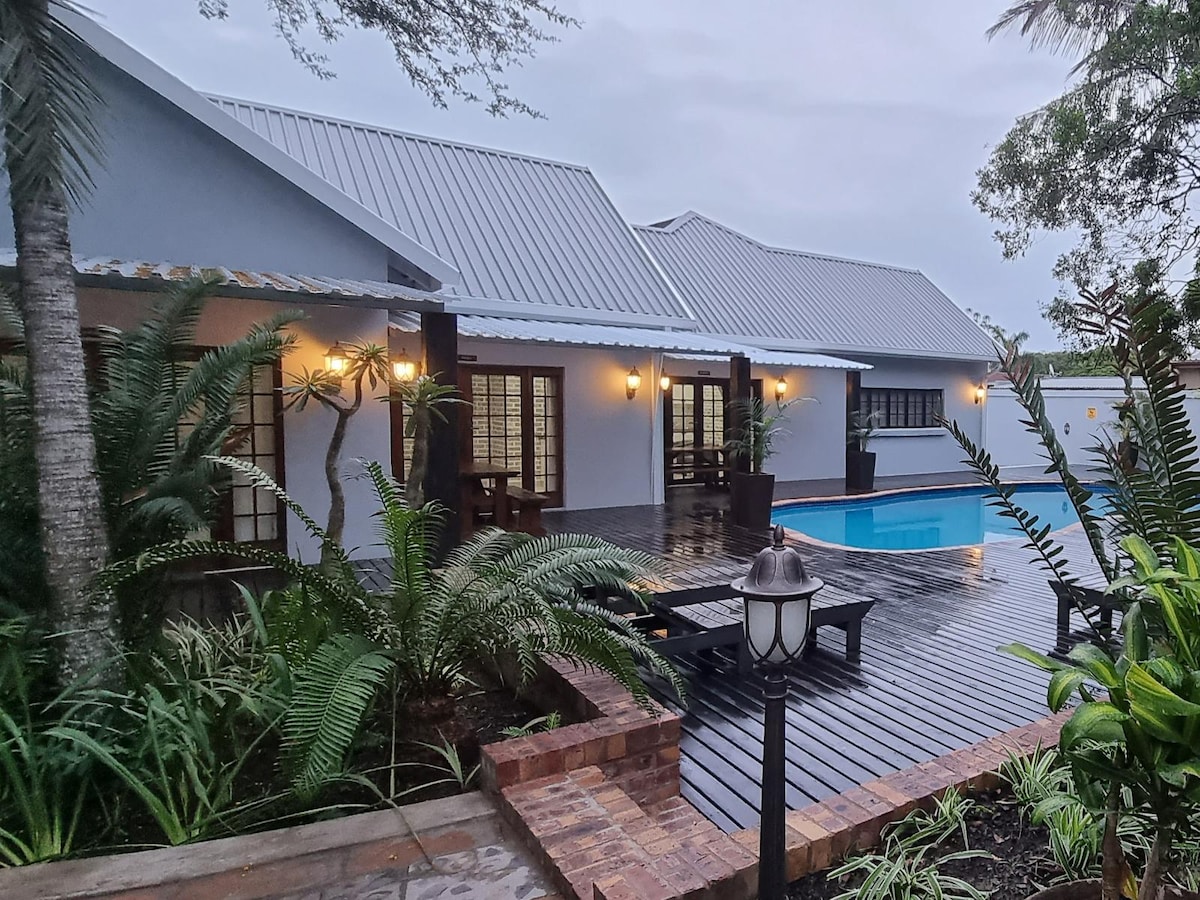 A spacious outdoor area features a wooden deck surrounding a pool, with lounge chairs arranged nearby. Lush greenery and palm trees add a tropical feel. The building showcases large windows and doors, enhancing the connection with the outdoor scenery.
