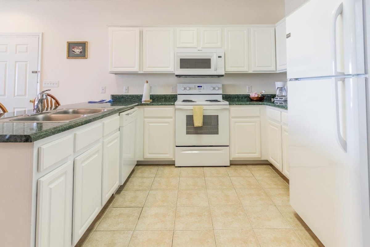 The kitchen features white cabinets and countertops, equipped with modern appliances including a microwave, stove, and refrigerator. A double sink is present, along with a small selection of cookware placed on the countertop. The tiled floor contributes to a clean and functional space.