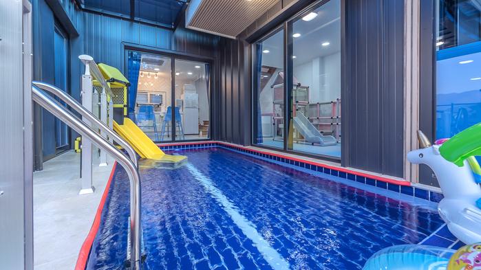 An indoor pool area features vibrant blue water, complemented by a bright yellow slide and a playful floating toy. Large glass windows provide views of the surrounding space, enhancing the openness of the area.