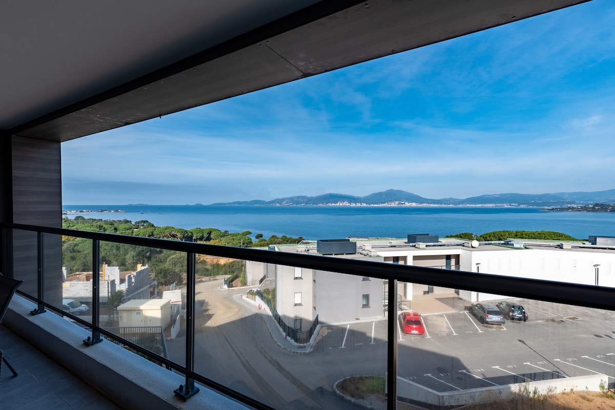 A panoramic view of the sea is framed by large glass windows, offering bright natural light and a sense of openness. The surrounding landscape includes green trees and several buildings along a calm coastline, showcasing the serene environment of Pietrosella.