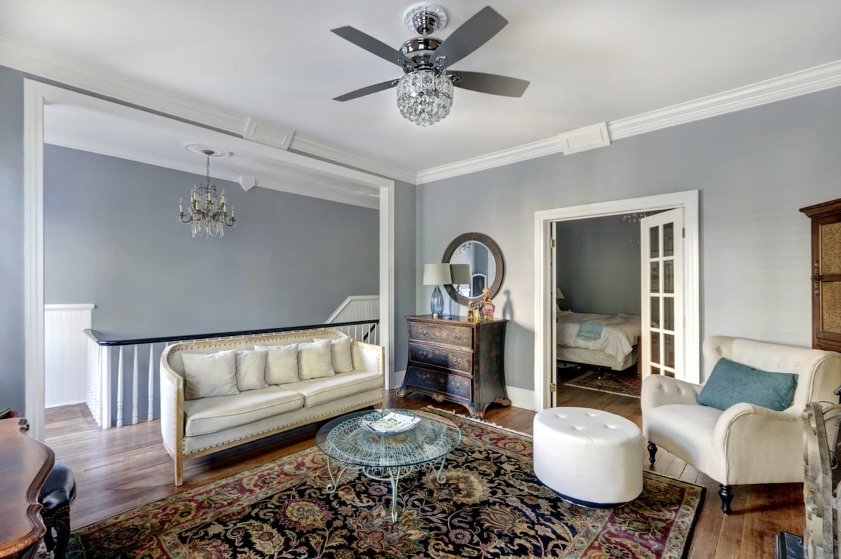 An open living area is showcased with hardwood flooring and light grey walls. Soft seating options include a white couch and an armchair. A decorative rug lies beneath a round coffee table. Elegant light fixtures are visible, and French doors lead to a bedroom space.