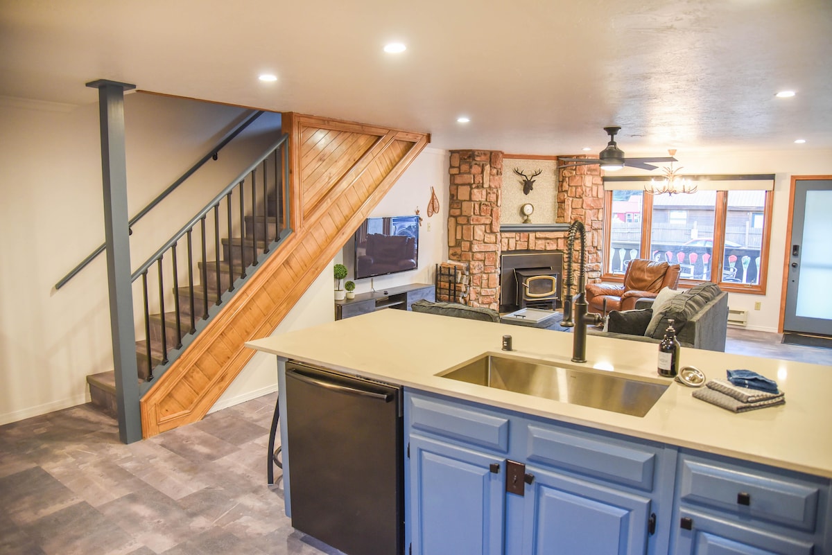 The open concept living area features a well-appointed kitchen with a large island and stainless steel appliances. A wood-burning stove is situated next to a stone fireplace, and the adjoining seating area includes a comfortable sectional couch. The wooden staircase leads to the upper levels.