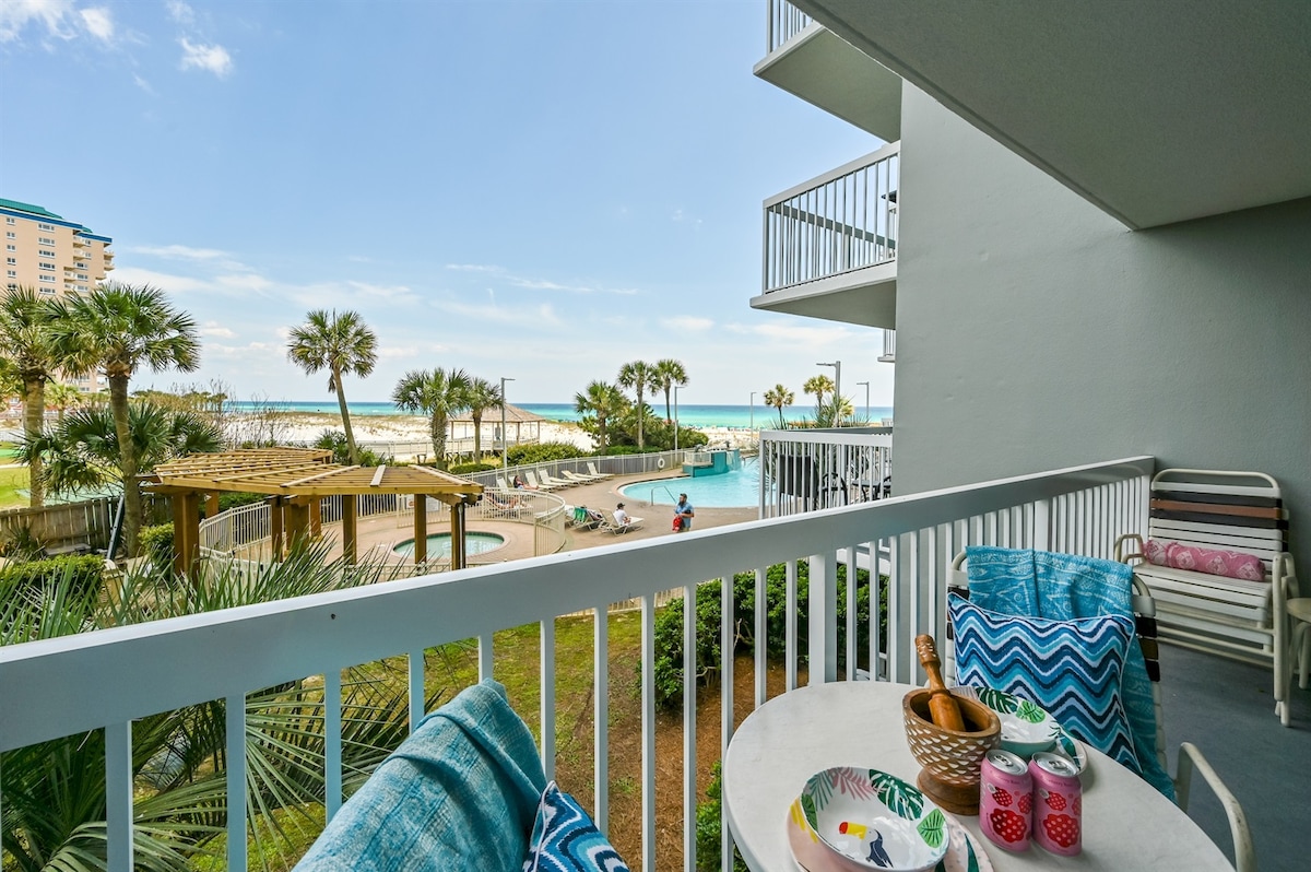 A spacious balcony features seating with colorful cushions, overlooking a lush landscape leading to the beach. A glimpse of the pool area and the Gulf is visible, framed by palm trees under a clear blue sky.