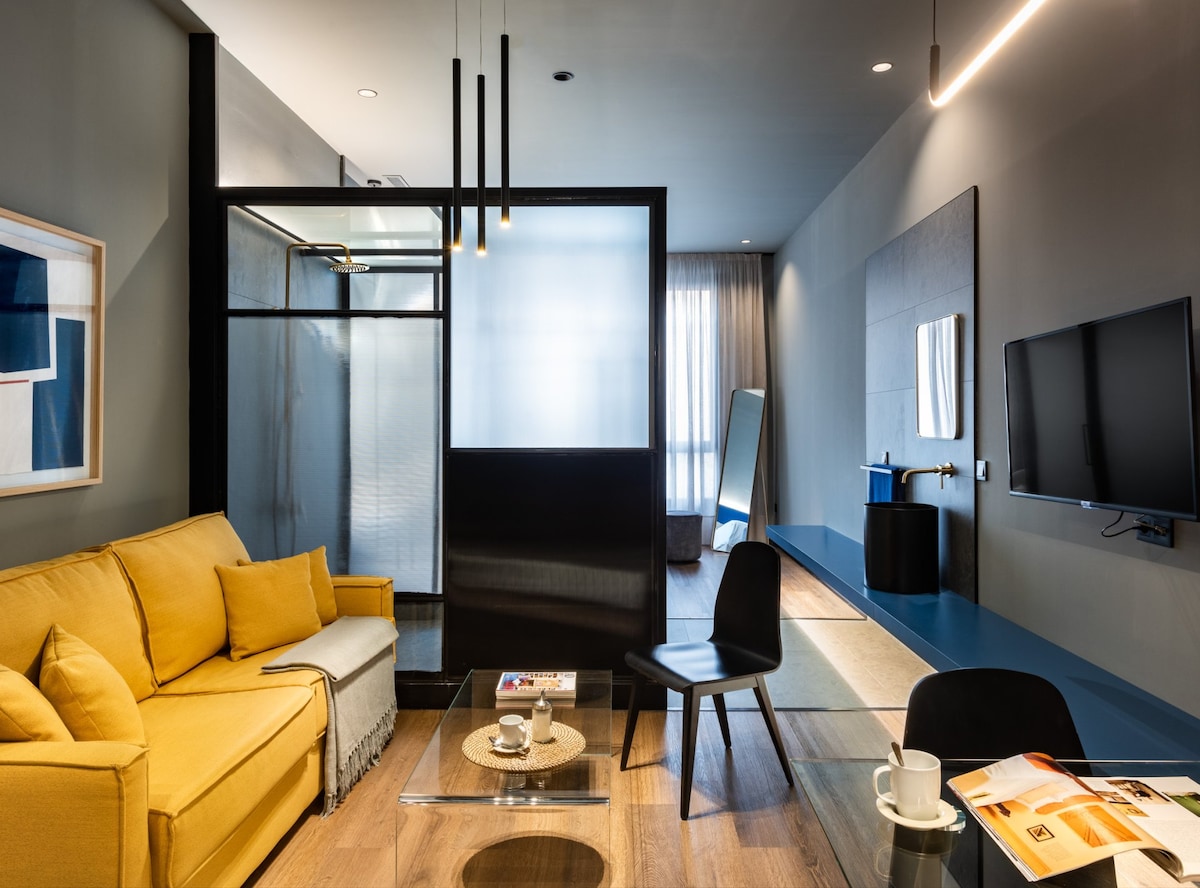 A modern living area is featured, showcasing a bright yellow sofa complemented by a glass coffee table. A black accent wall provides contrast, while a spacious window allows natural light to illuminate the room. A television is mounted on the wall opposite a sleek mirror.