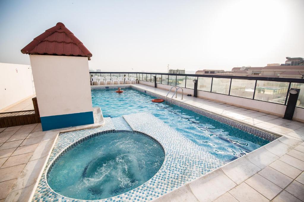 A rooftop pool area features a rectangular pool beside a circular hot tub. The pool is surrounded by tiled flooring and framed by a glass railing, offering an open view of the surroundings. A shaded structure with a tiled roof is positioned nearby.