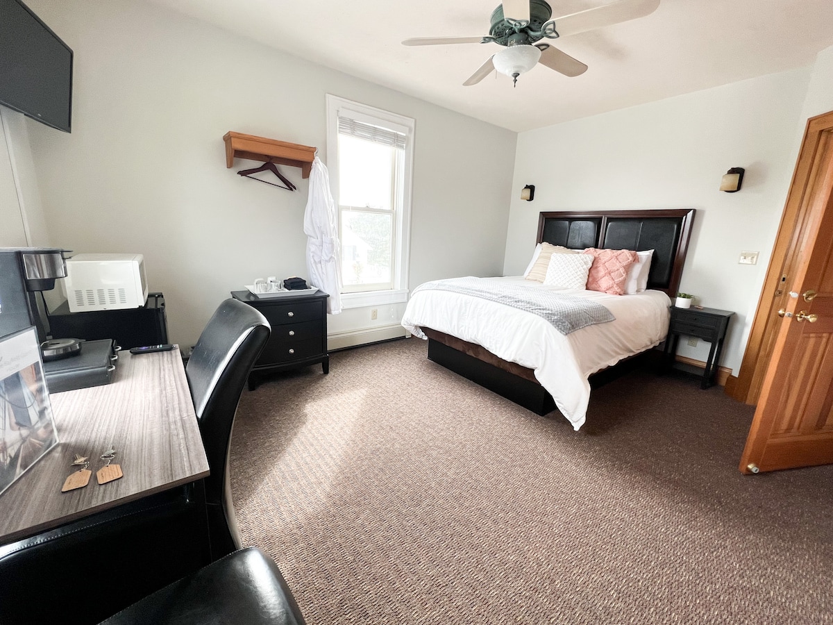 The spacious queen suite features a comfortable bed with decorative pillows, a desk with a chair, and a dresser. Natural light enters through the window, and a plush rug covers the floor. A television is mounted on the wall, adding convenience to the room.