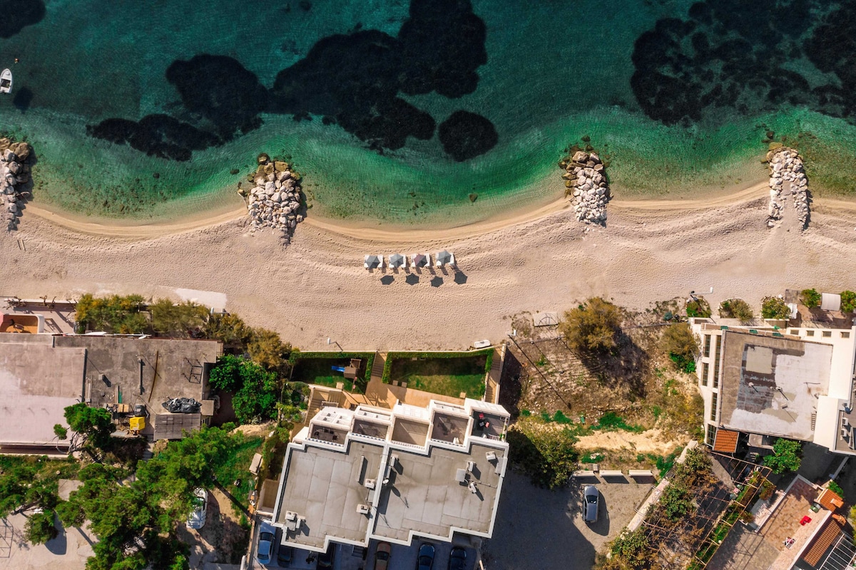 An aerial view captures the pristine beach bordered by gentle waves. Sun umbrellas and loungers align along the shoreline, contrasting the golden sand. Coastal vegetation and buildings are visible, providing a glimpse of the local landscape surrounding the tranquil waters.