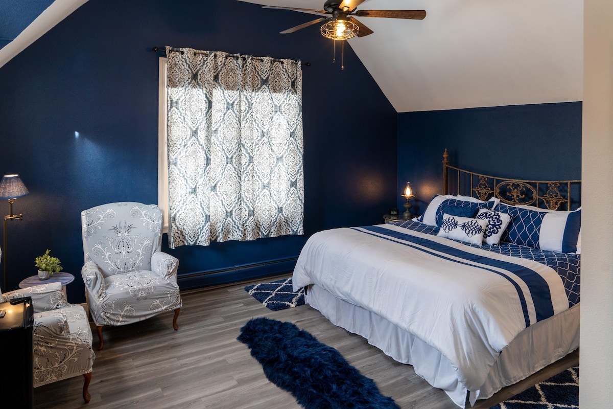 A spacious bedroom features deep blue walls and light gray accented furnishings. A large bed with crisp white linens and blue decorative pillows is complemented by a plush area rug. Natural light enters through a large window with patterned curtains. A cozy reading chair adds to the comfort.