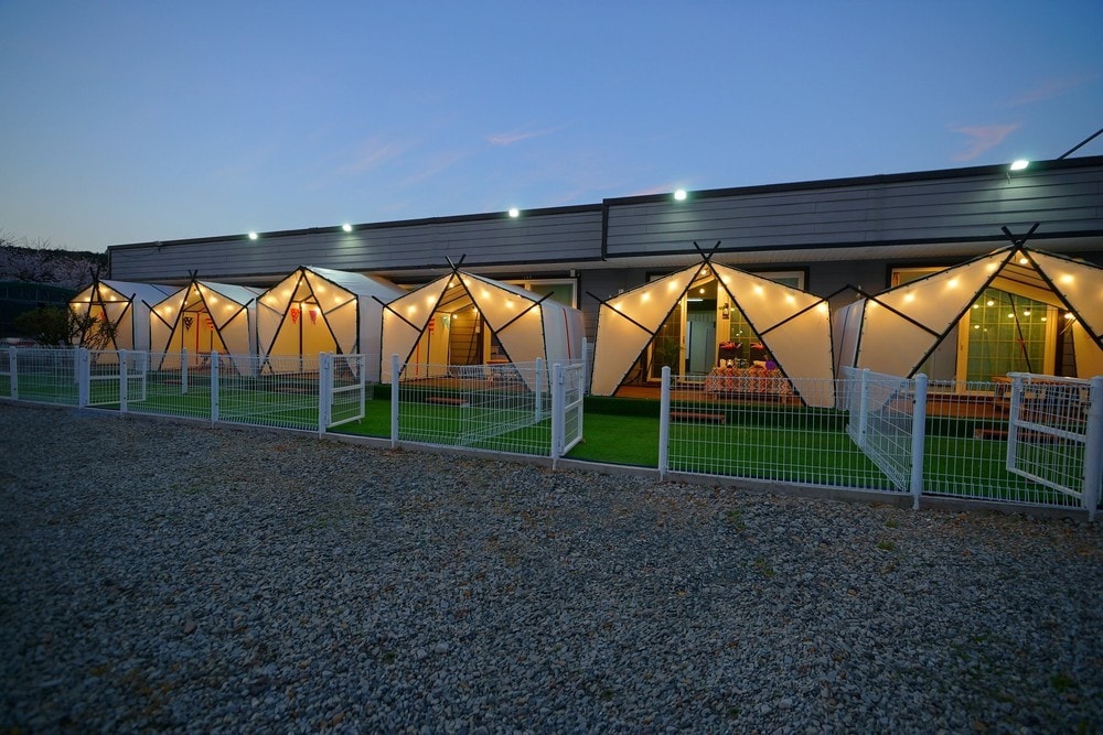 A row of modern tents with illuminated entrances is visible, set against a twilight sky. Each tent features a distinctive fabric design and is positioned on a grassy area. A white fence surrounds the space, providing a cozy and secure environment.