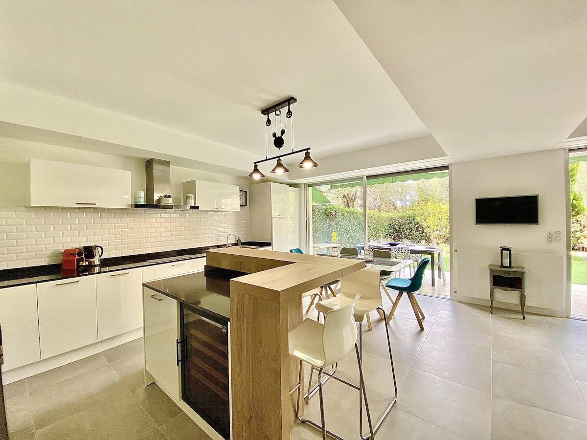A modern open kitchen features sleek cabinetry and a large island with seating for four. Natural light streams in through expansive glass doors that lead to the garden terrace. Appliances, including a coffee maker and wine cooler, are positioned for convenience in the spacious area.