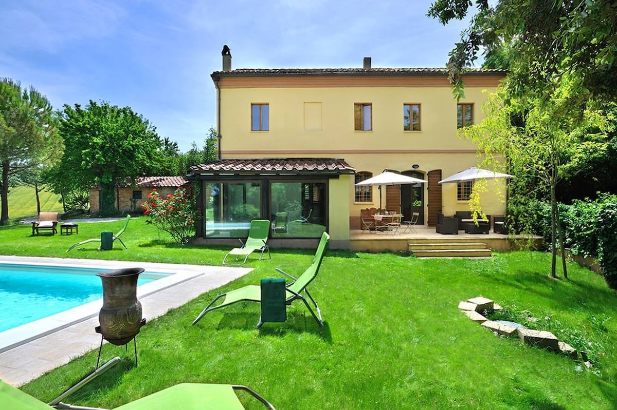 A villa is set amidst lush greenery, featuring a private pool surrounded by lounge chairs. A spacious patio with dining areas is visible alongside the pool, while large glass windows reflect natural light. The exterior blends seamlessly with its tranquil surroundings.