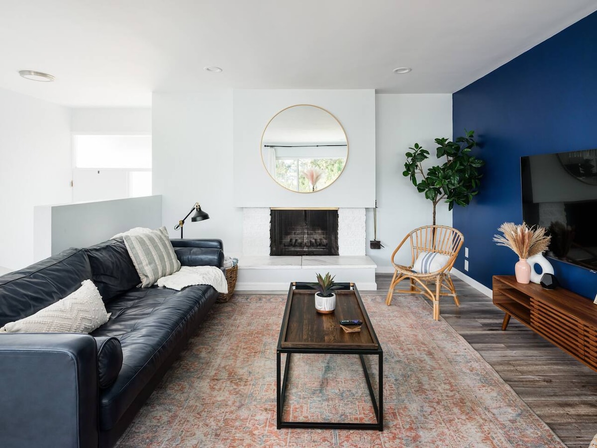 A spacious living room features a black leather sofa and a wooden coffee table atop a patterned rug. A large round mirror is centered above a chic white fireplace. Natural light fills the room, highlighting a green plant by the wall and a TV on an elegant media console.