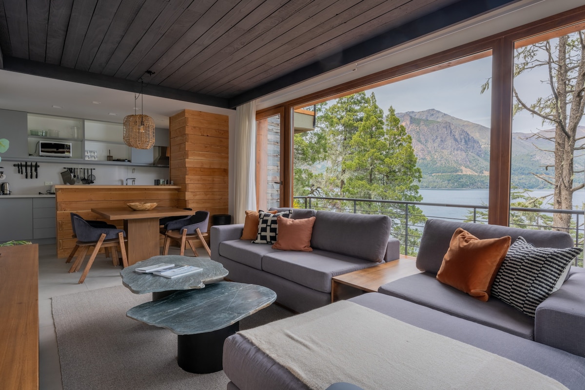 A modern living area features large windows that frame a view of the lake and mountains. Gray sofas are arranged around a unique coffee table. A wooden dining table is visible nearby, complemented by natural materials in the design.