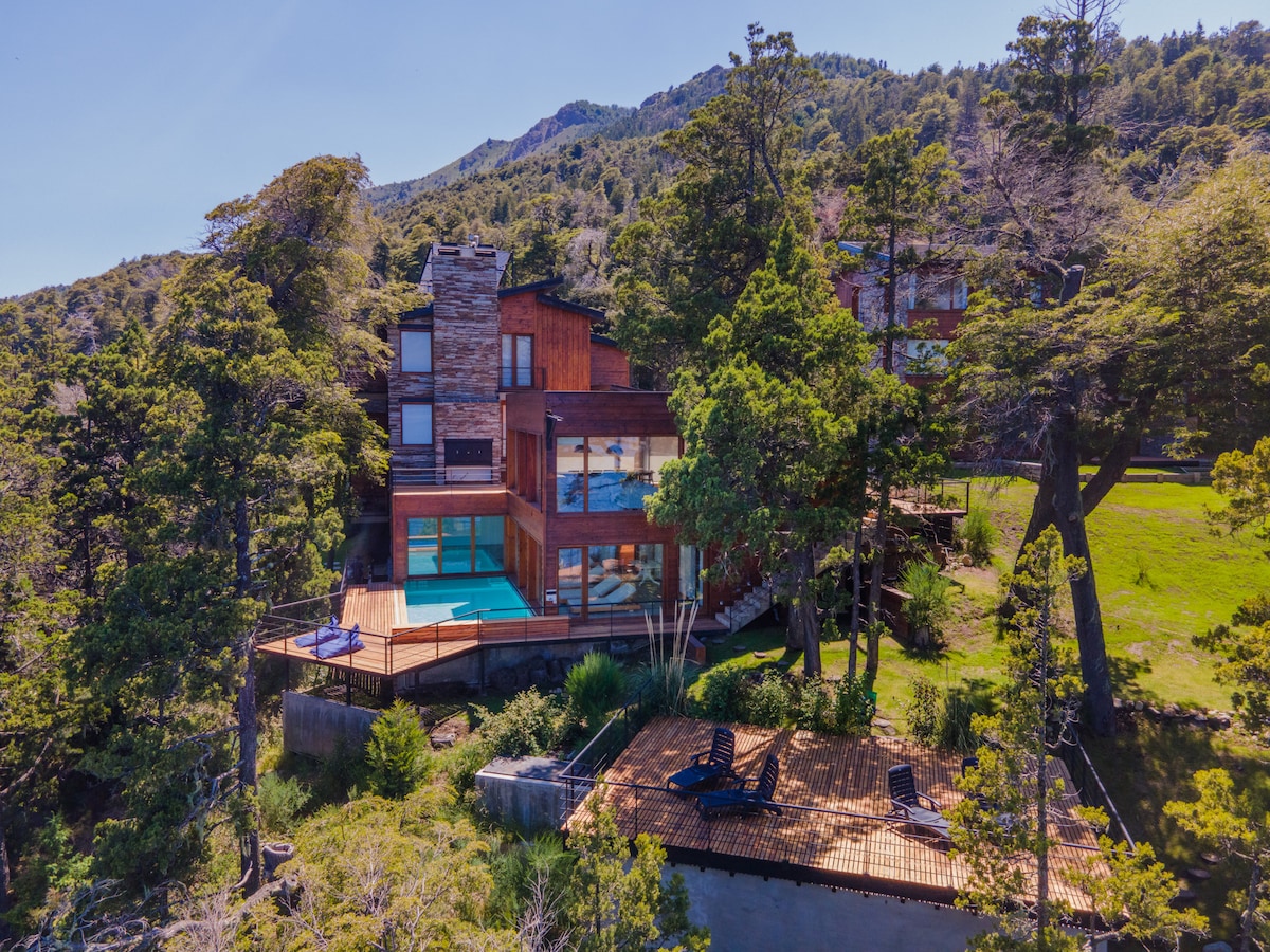 A spacious apartment exterior is framed by lush greenery and trees, featuring large glass windows that reflect the natural surroundings. An outdoor deck with seating and a pool offers a serene spot for relaxation, while mountains rise in the background, highlighting the picturesque setting.