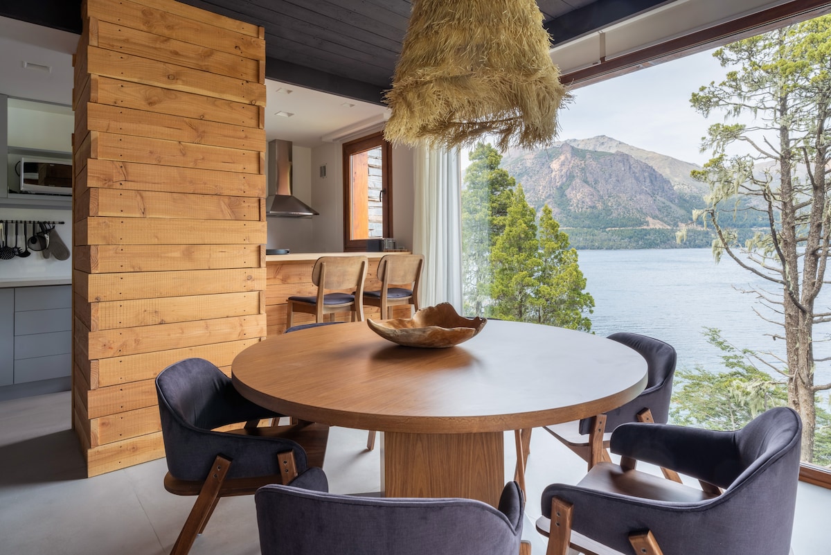 A round wooden dining table is surrounded by four chairs, set against large glass doors offering views of Gutiérrez Lake and the surrounding mountains. A pendant light made of natural materials hangs from the ceiling, adding an organic touch to the space.