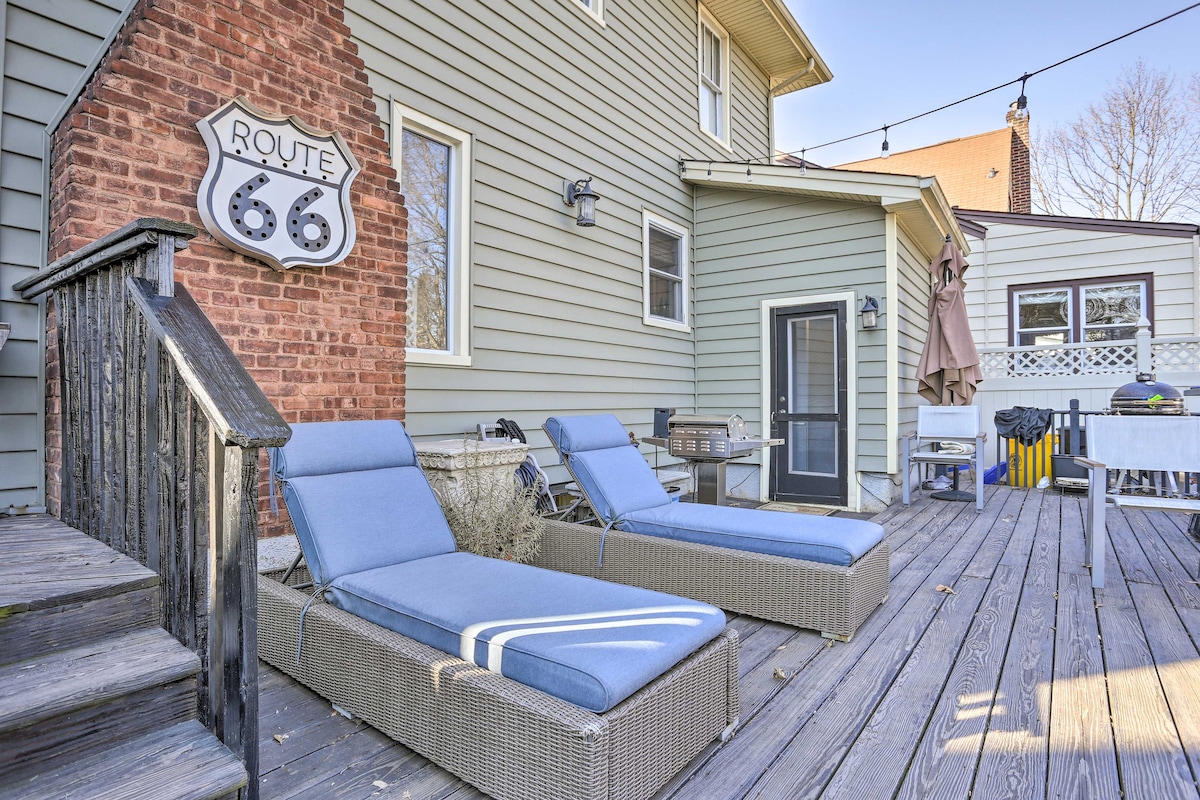 A wooden deck is highlighted by two lounge chairs positioned for relaxation. A charcoal grill is visible nearby, along with a dining area sheltered by an umbrella. The exterior wall features a decorative Route 66 sign, enhancing the outdoor space.