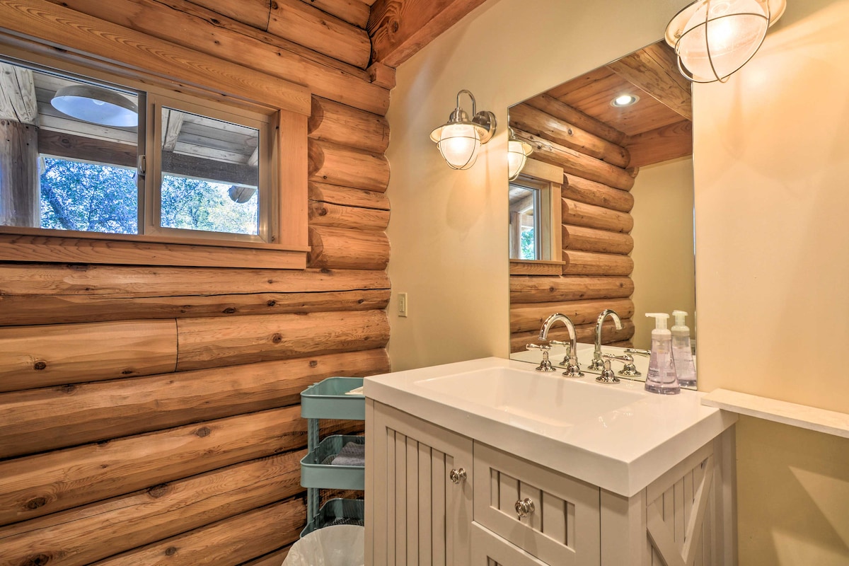 The bathroom features natural log walls, enhancing the cabin's rustic charm. A sleek white vanity with a dual faucet sink is accented by modern lighting above the mirrors. A window allows natural light to fill the space, while a small cart provides additional storage.