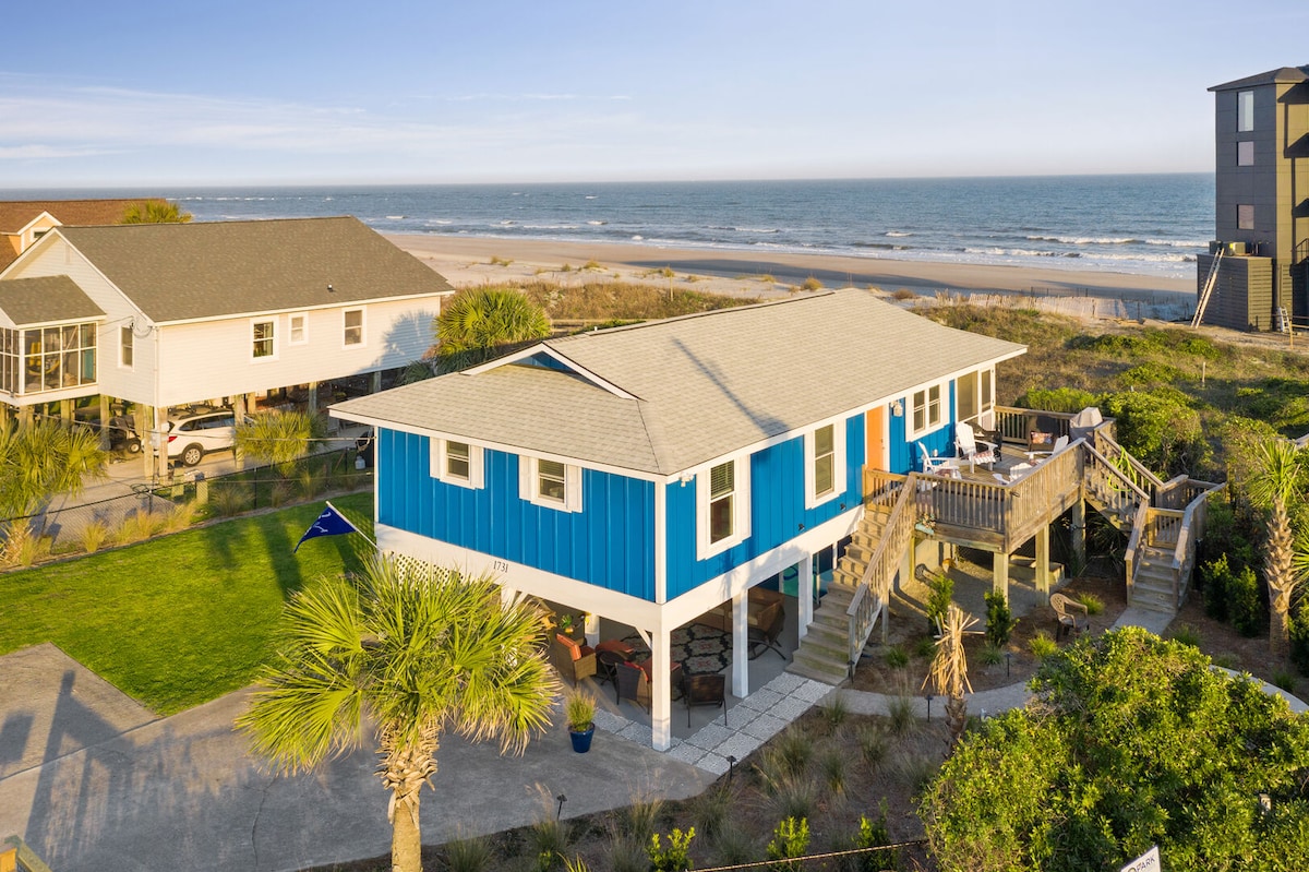 A coastal cottage is prominently featured, with a vibrant blue exterior and a spacious deck overlooking the ocean. Lush greenery and palm trees are present in the yard, while the beach is visible in the background, enhancing the connection to the seaside setting.
