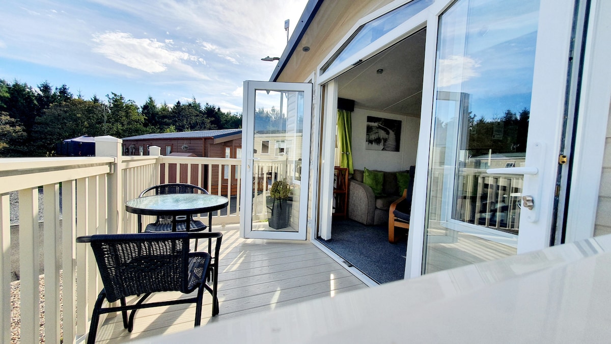 A private balcony area features a round glass table accompanied by two black wicker chairs. Floor-to-ceiling doors open to a living space, allowing natural light to fill the room. Green foliage can be seen in the background, adding a hint of nature.