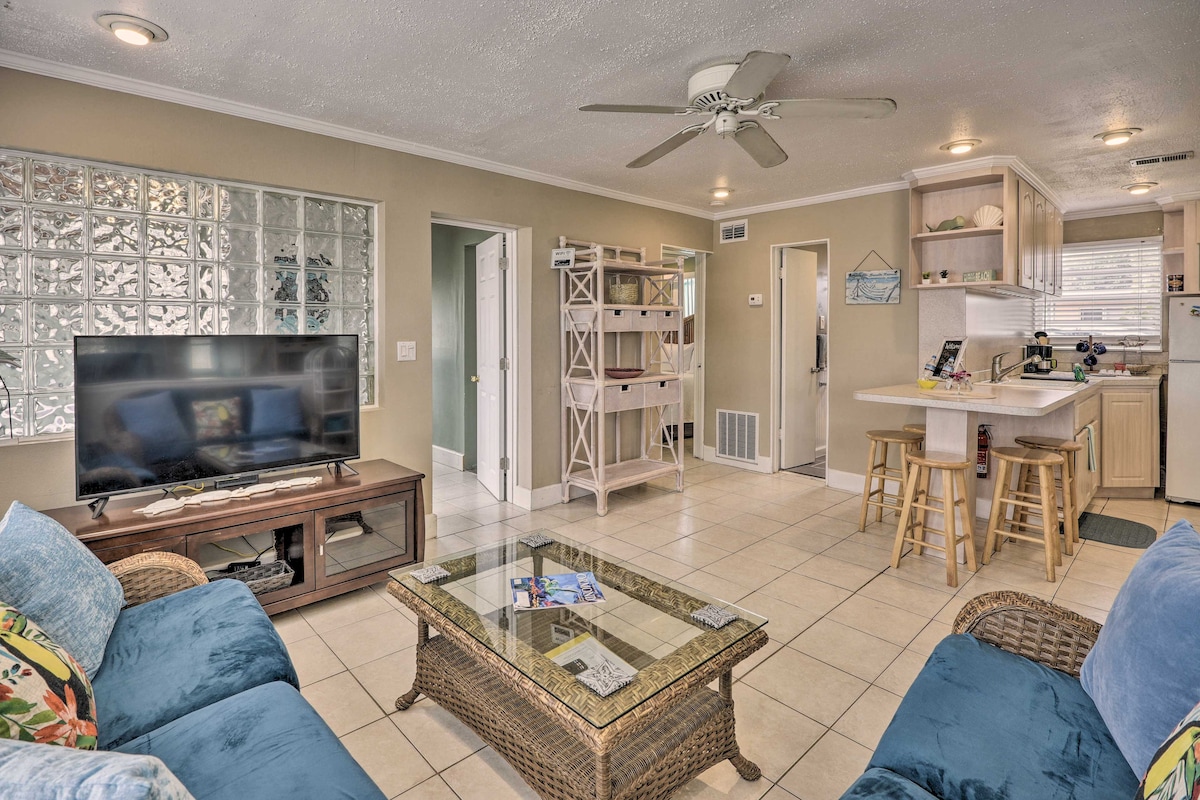 An open living area is showcased, featuring tile flooring and large windows allowing natural light. A ceiling fan is present above the seating area, which includes a blue sofa and a coffee table. A Smart TV sits on a console, complemented by a kitchen with bar seating.