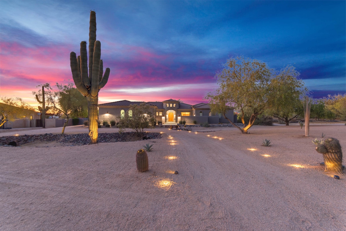 The exterior of a desert home is featured during twilight, with a colorful sunset illuminating the sky. Pathway lights guide the view towards the entrance, surrounded by desert flora, including tall cacti and other native plants, enhancing the tranquil ambiance.