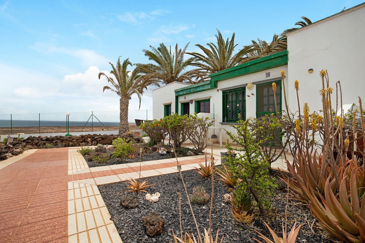 The exterior of the holiday apartment features a landscaped garden with various plants and stone paths. Tall palm trees provide shade and a view of the sea can be seen in the background. The white building is accented with green trim, creating a fresh appearance.