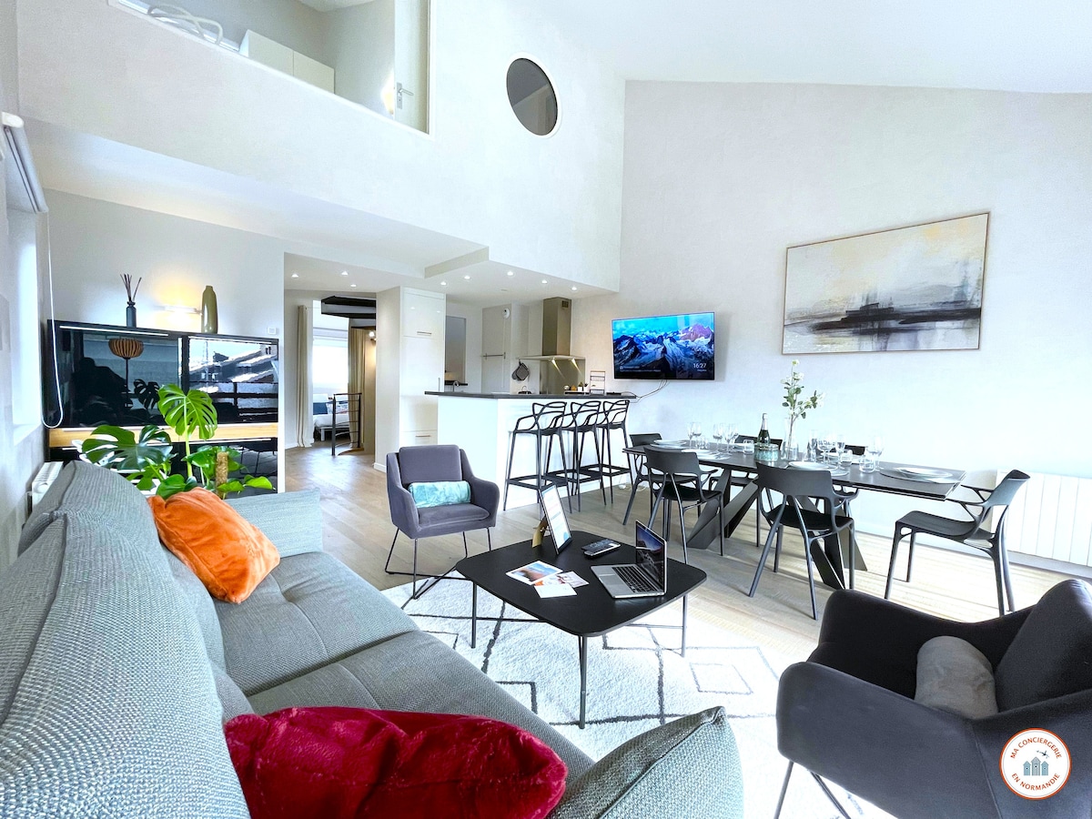 A spacious living area is prominently featured, showcasing a comfortable sofa adorned with colorful cushions. A modern dining table with black chairs is positioned nearby, while a Smart TV is mounted on the wall. Soft lighting enhances the contemporary décor, complemented by a well-equipped kitchen area in the background.