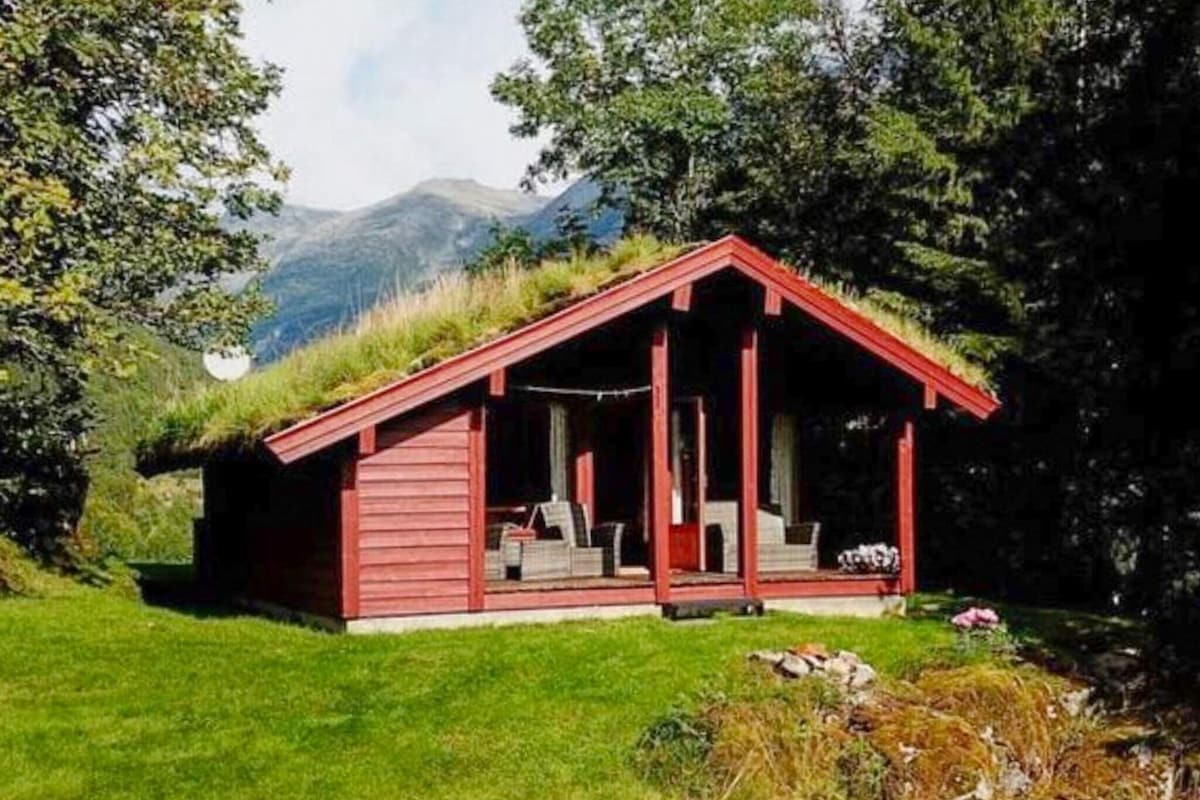 A charming red cabin features a green grass roof and is surrounded by trees and grassy areas. Outdoor seating is provided on a covered porch, with mountain views visible in the background. Sunlight gently illuminates the structure, creating a peaceful retreat.