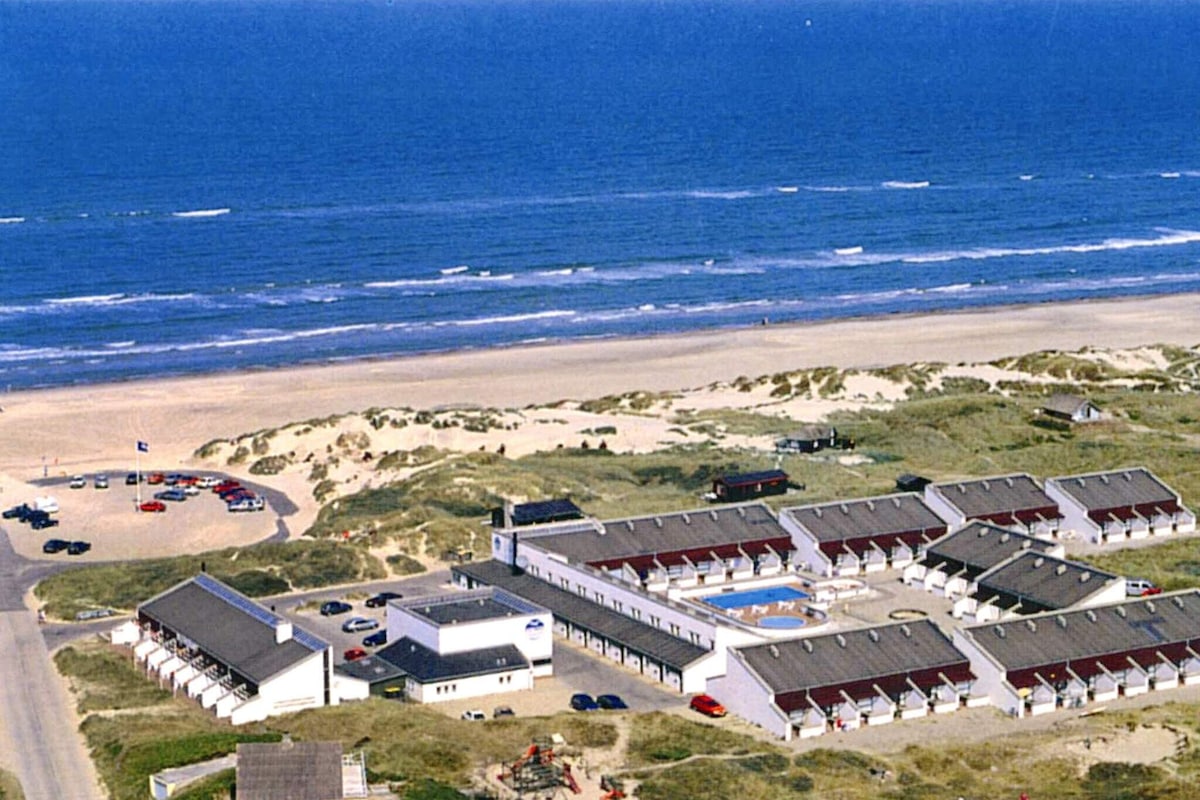 An aerial view captures a holiday home complex adjacent to a sandy beach, with the North Sea visible in the background. The scene includes rows of holiday homes, a shared swimming pool area, and a nearby parking space. Waves lap gently against the shore.