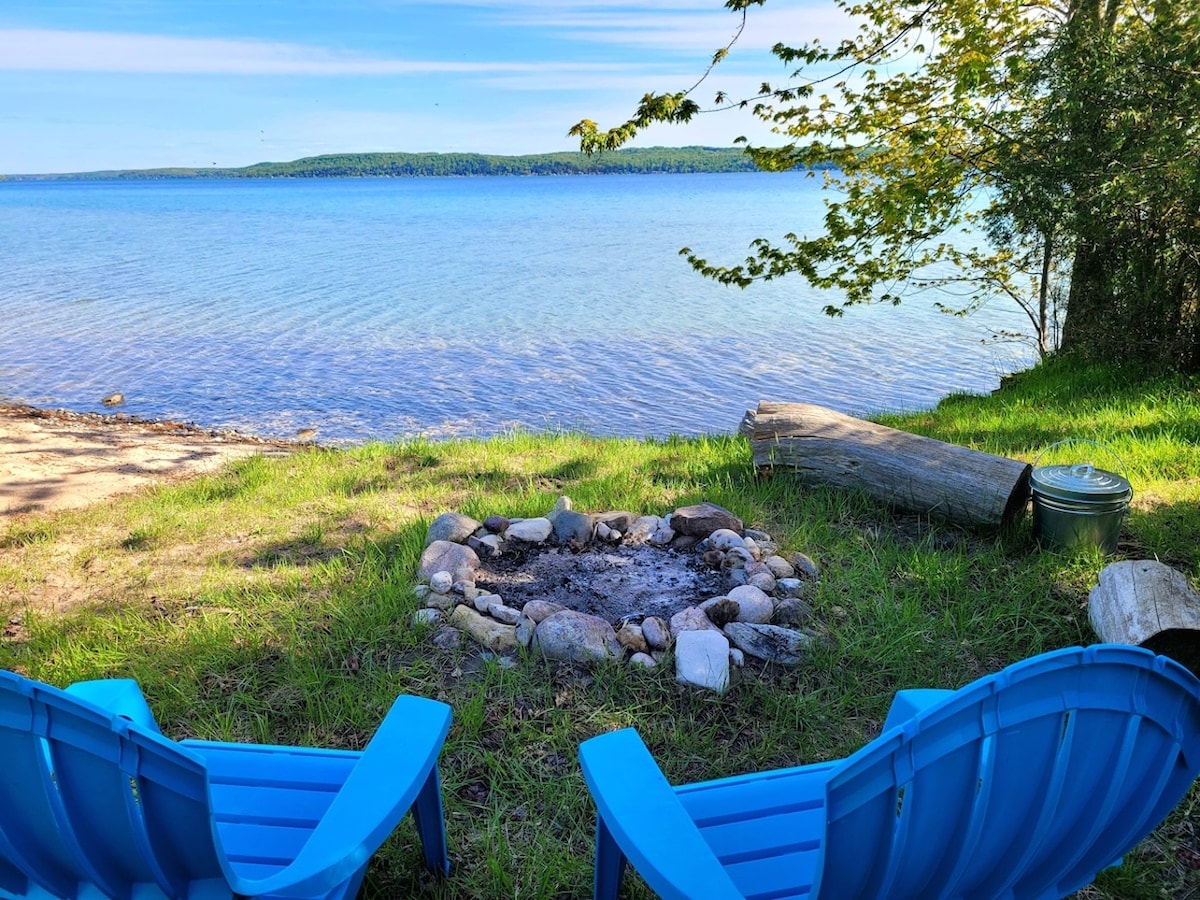 Two blue Adirondack chairs face a serene waterfront with clear, calm waters. A small bonfire pit made of stones is positioned nearby, surrounded by grassy areas and a few logs. The scene is framed by trees, offering a peaceful outdoor setting.
