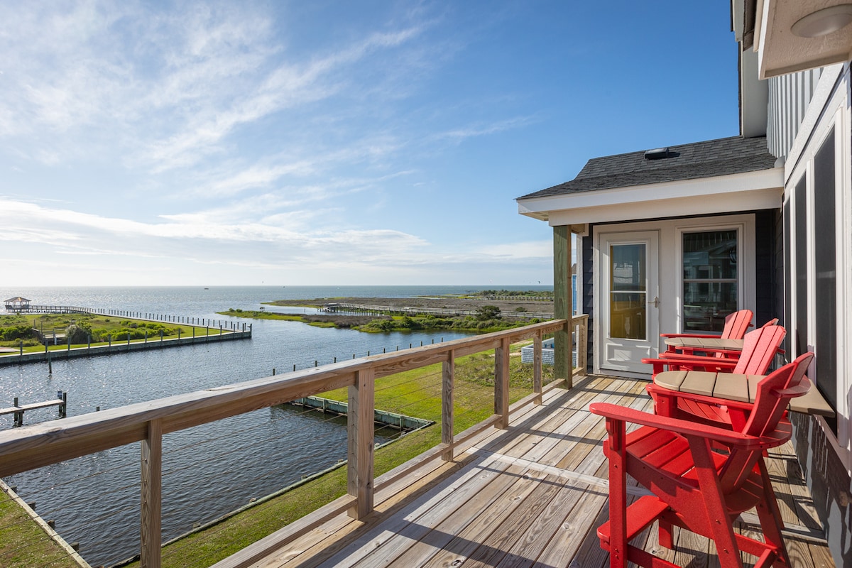 A wooden deck features two vibrant red Adirondack chairs overlooking the calm waters of Pamlico Sound. Lush green grass and a clear sky create a tranquil backdrop, while the expansive view includes a distant pier stretching into the horizon.