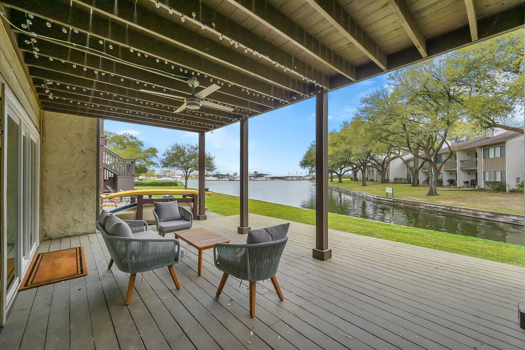 An outdoor seating area features comfortable chairs surrounding a wooden table. The space overlooks a peaceful canal lined with trees, creating a calm atmosphere. Twinkling string lights hang from the ceiling, adding subtle charm, and expansive decking extends for additional outdoor enjoyment.
