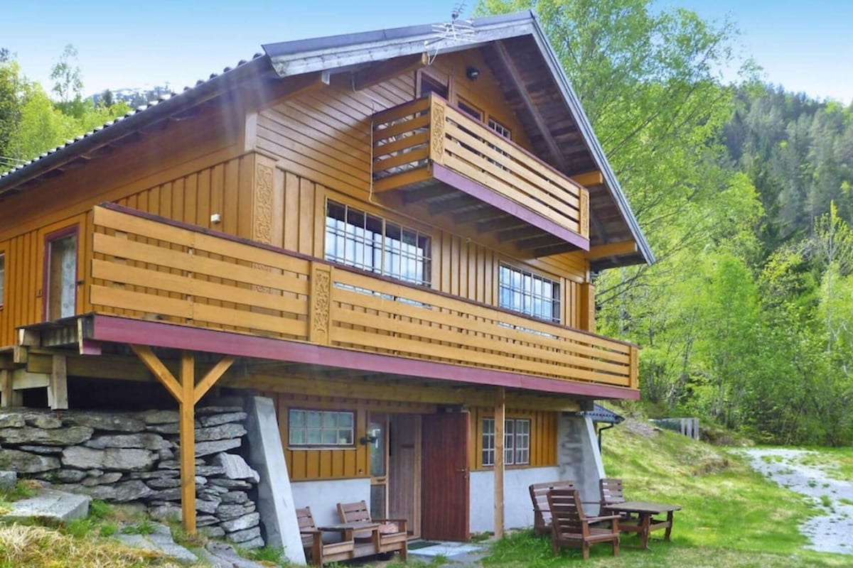 A two-story wooden holiday home is shown, featuring a balcony with a wooden railing. The structure is supported by stone pillars, with a small porch and outdoor seating area visible in front. Surrounded by greenery, the setting offers a natural, serene backdrop.